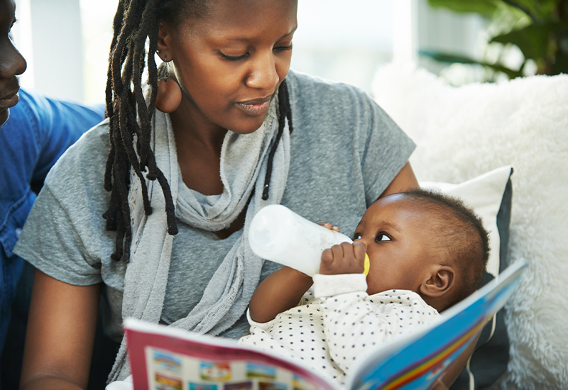 Mother holding baby with bottle and book in other hand on couch