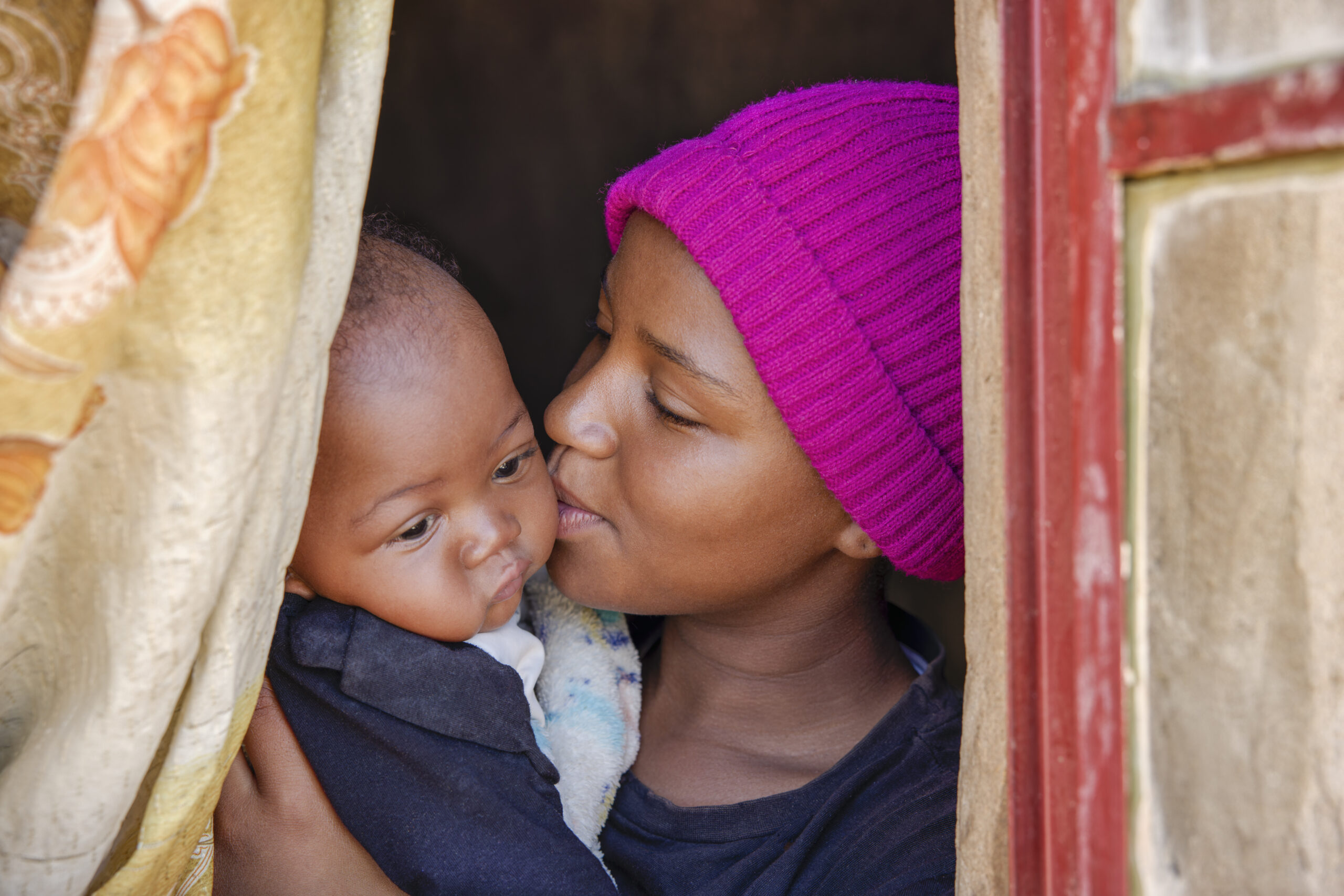 Mom in pink hat kissing babies cheek near paint peeling window