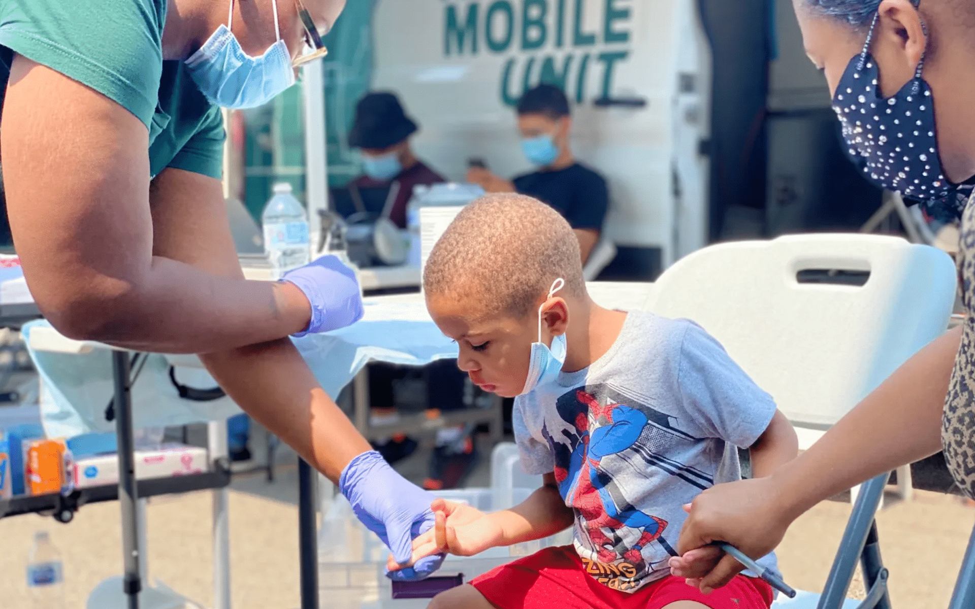 Young boy getting a blood finger prick test sitting next to his mom holding a clipboard